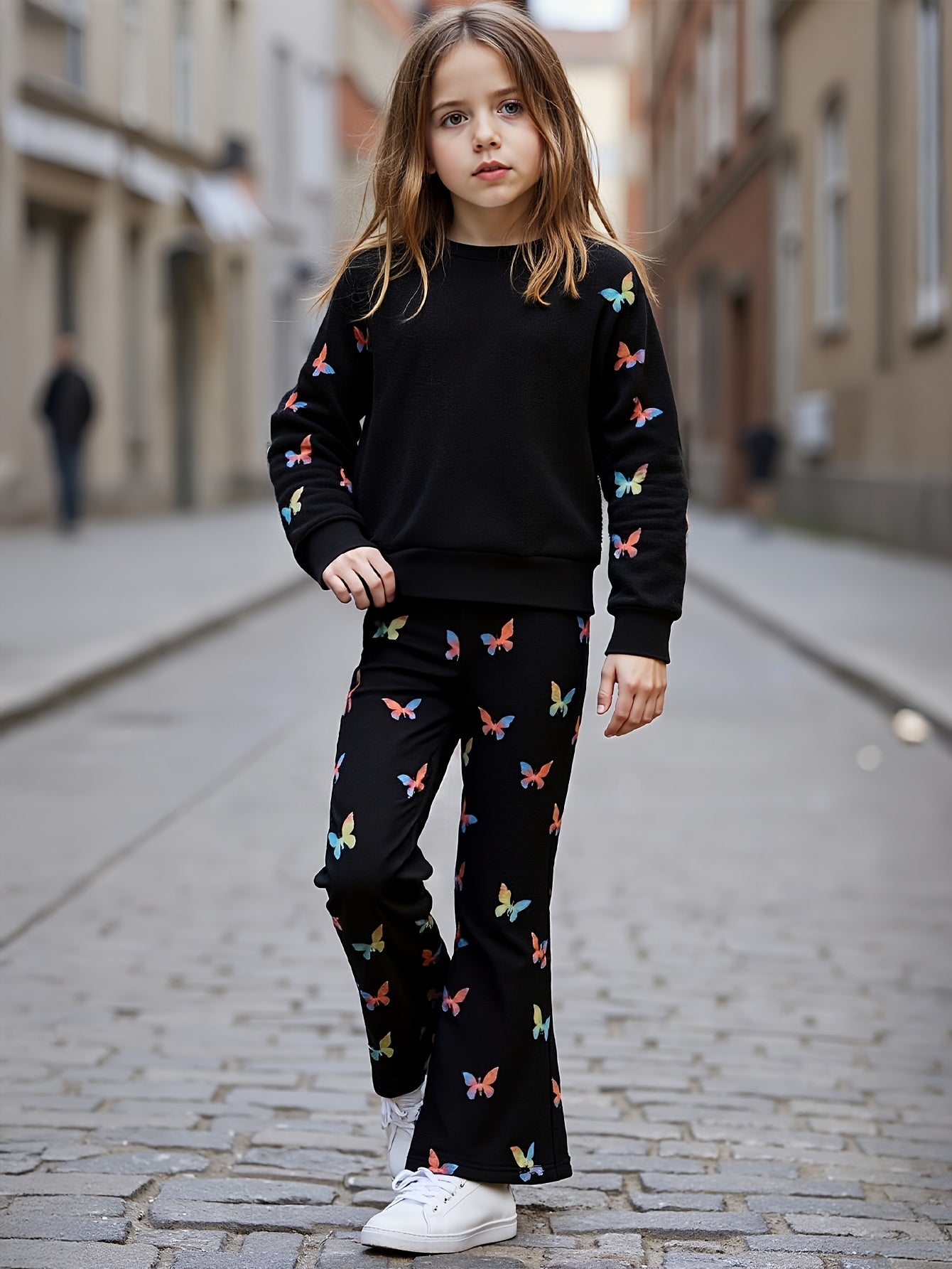 A young girl stands on a cobblestone street wearing a matching black sweatshirt and flared pants set, both covered in a colorful butterfly print. She is wearing white sneakers.