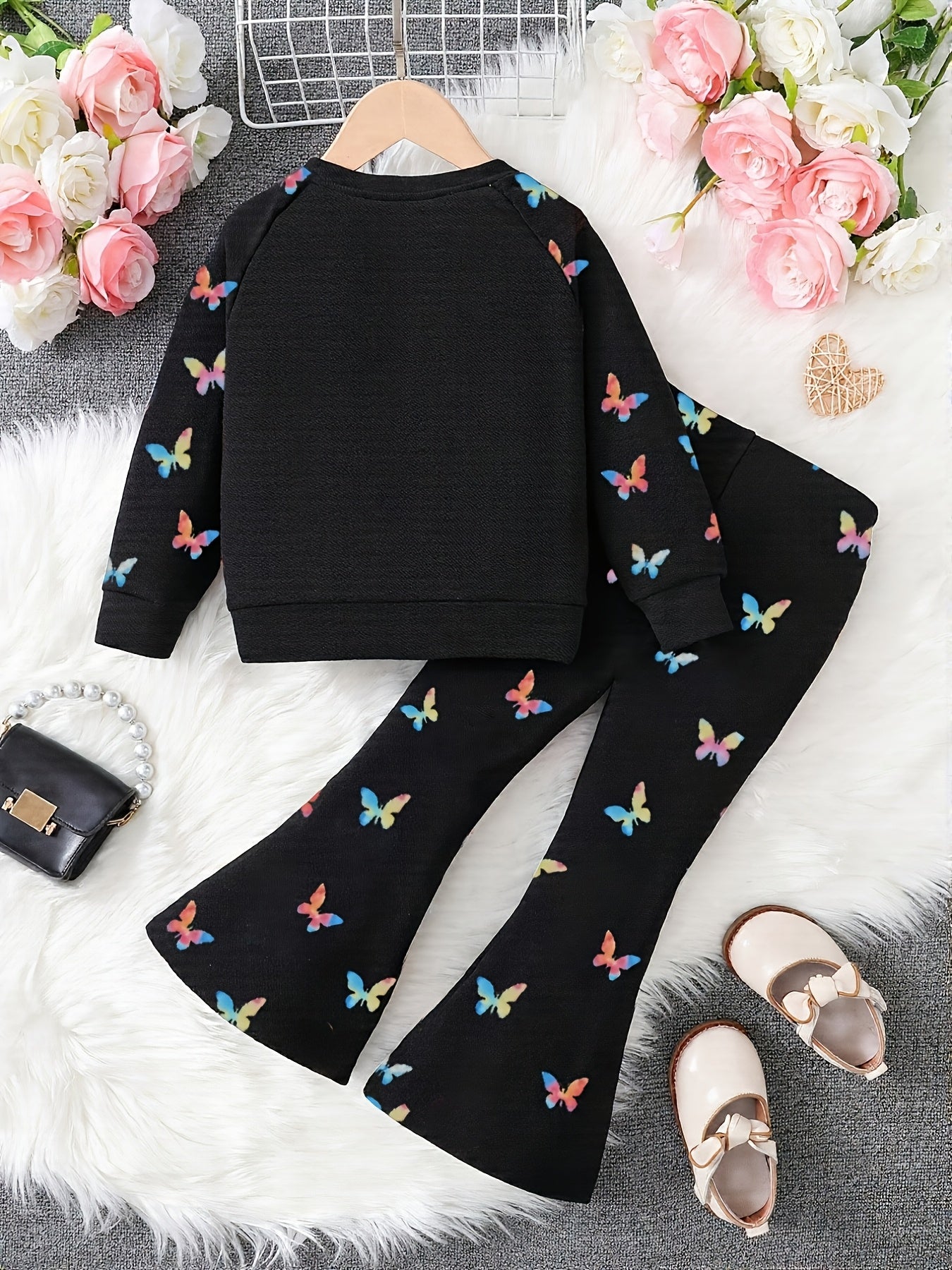 A two-piece children's outfit set laid flat on a white faux-fur surface, featuring a black long-sleeve top and matching black flared pants, both printed with small multicolored butterfly motifs. The set is styled with pink and white roses, a small black purse, and beige shoes.