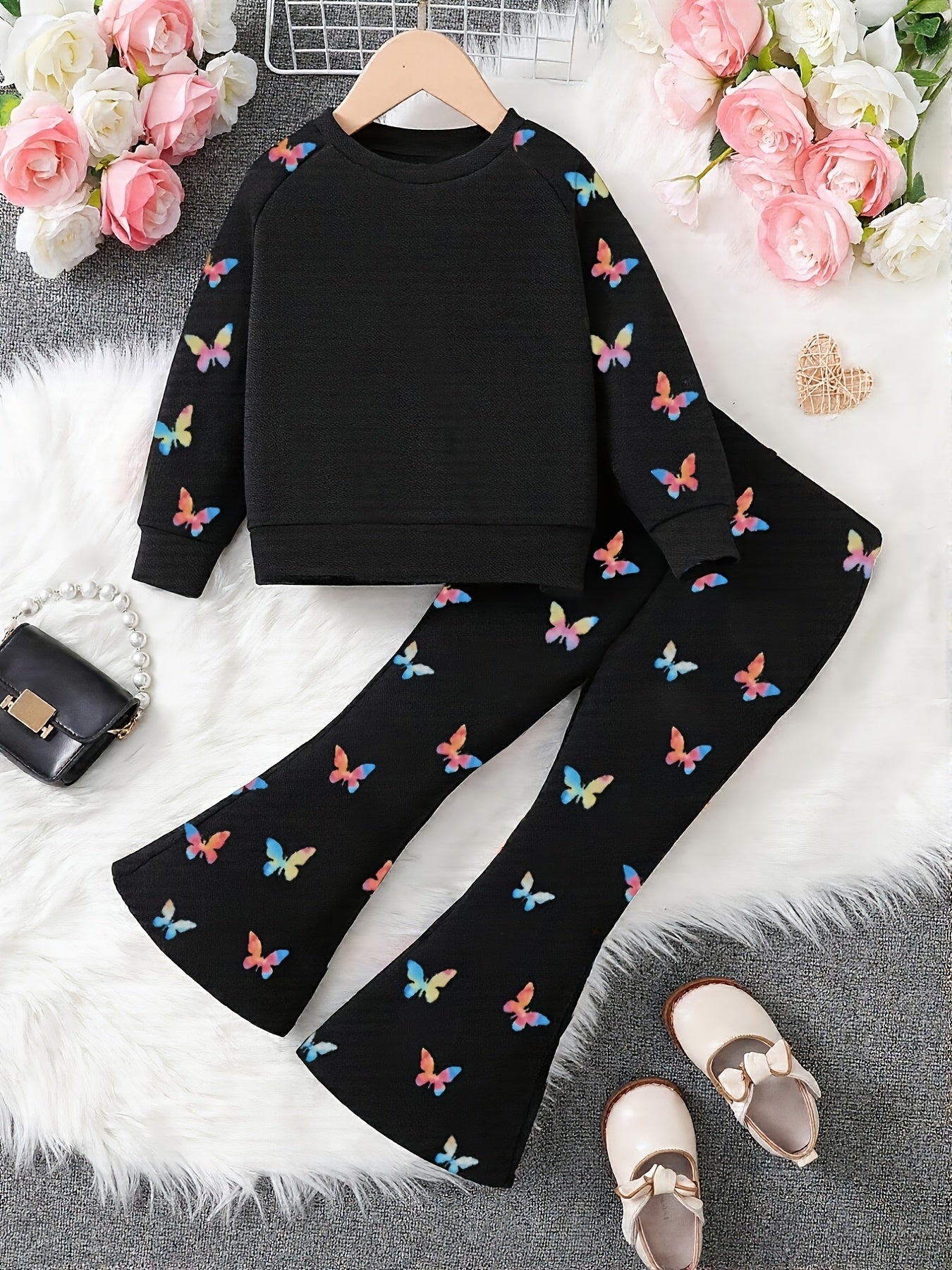 A children's two-piece outfit laid flat on a white faux-fur surface, featuring a black long-sleeve top and matching black flared pants with colorful butterfly prints. The set is styled with pink flowers, a small black purse, and beige shoes.