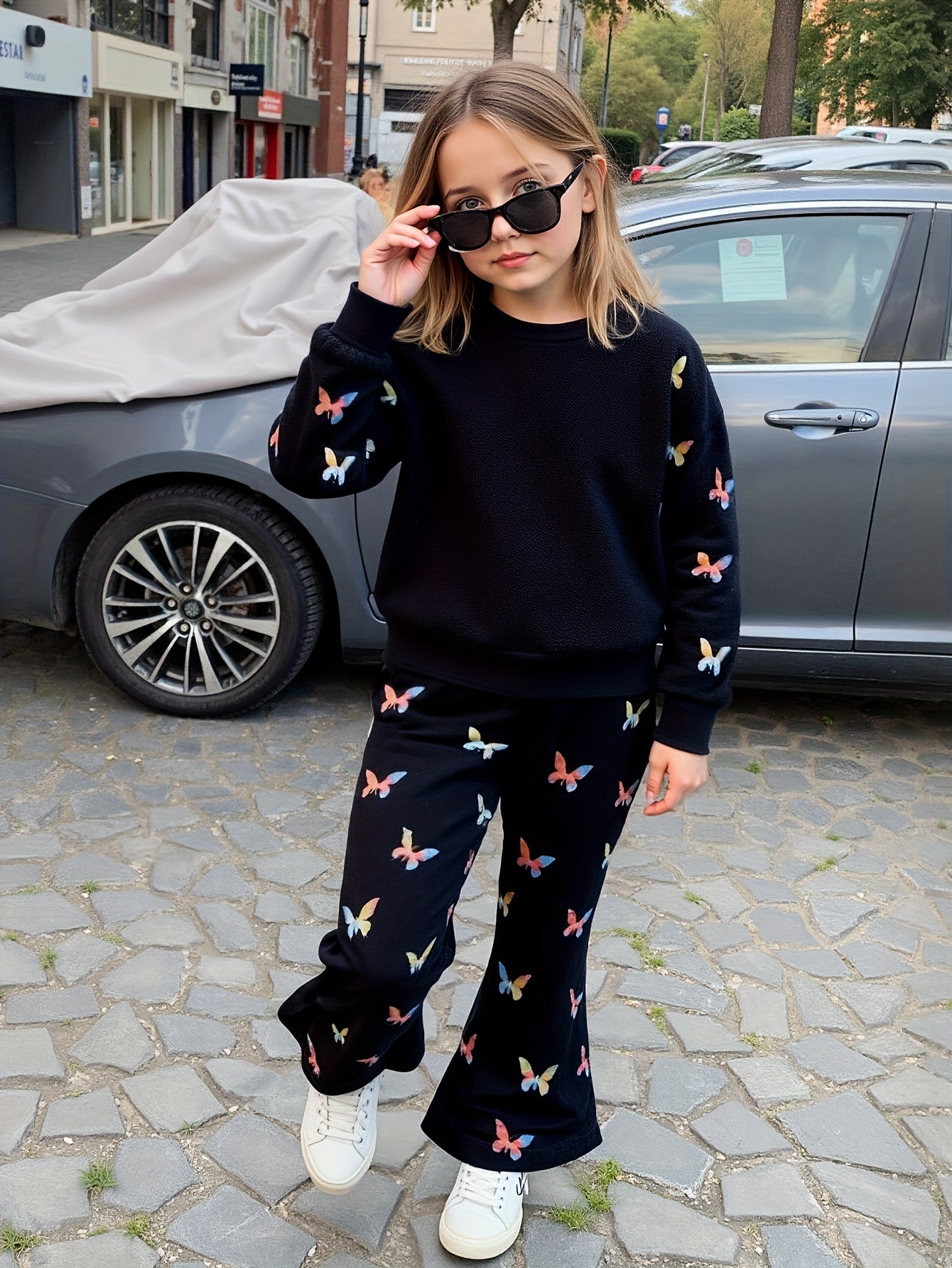 A young girl wearing a matching black sweatshirt and wide-leg pants set with a colorful butterfly print, styled with black sunglasses and white sneakers, standing on a cobblestone street in front of a gray car.