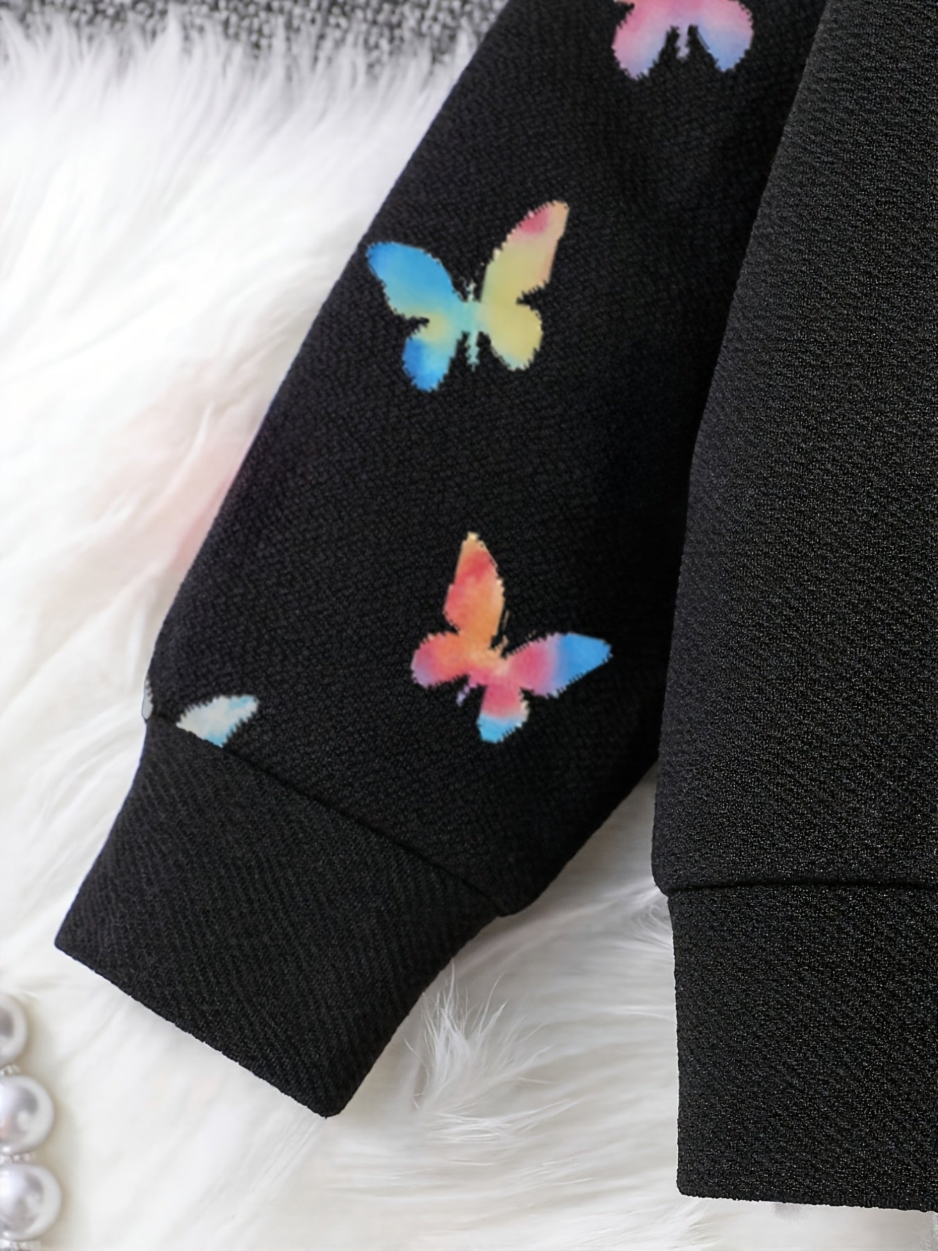 Close-up of a black ribbed sweatshirt sleeve decorated with colorful butterfly prints in shades of blue, yellow, pink, and orange, resting on a white faux fur surface.