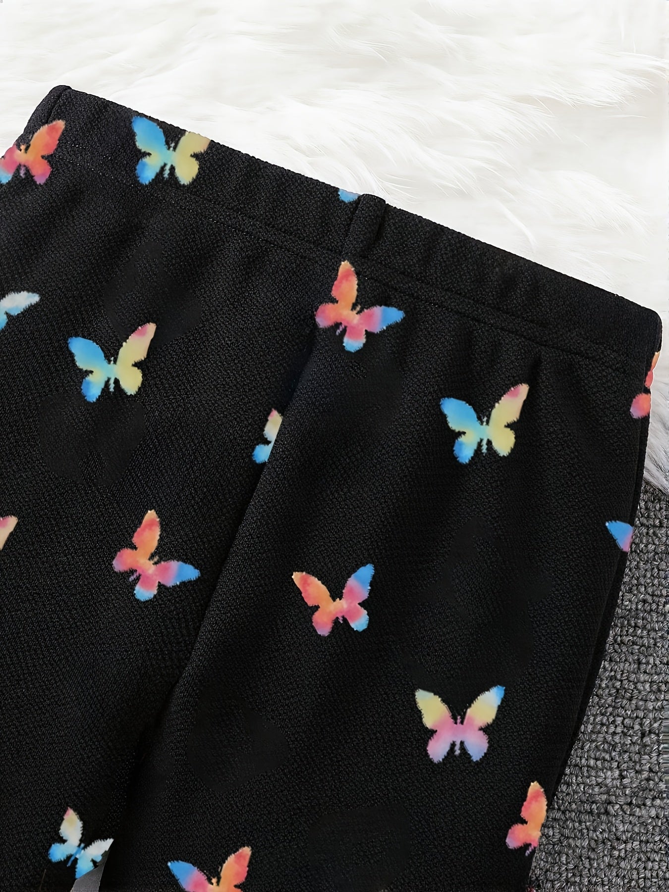 Black textured knit pants featuring an all-over colorful butterfly print, laid flat on a white faux fur surface.