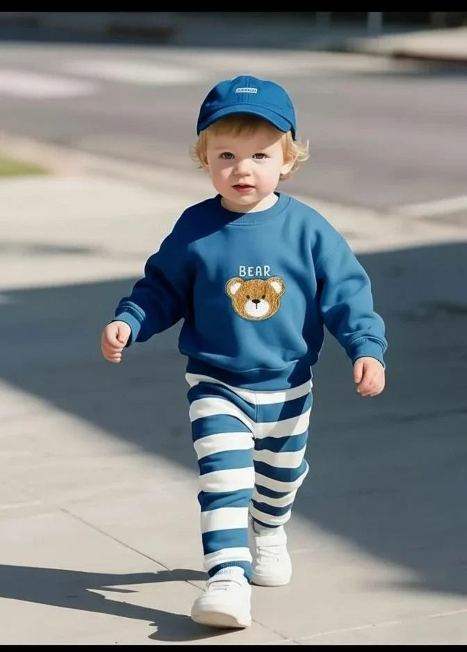 A toddler walking outdoors wearing a blue sweatshirt with a teddy bear graphic, blue cap, blue and white striped pants, and white sneakers.