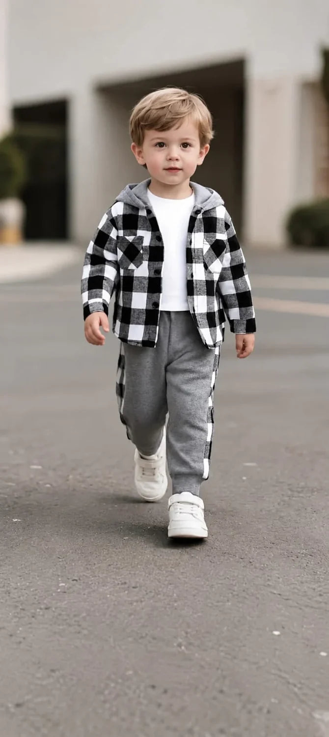 A toddler boy walking outdoors wearing a black and white buffalo plaid hooded flannel shirt over a white t-shirt, paired with gray sweatpants and white sneakers.