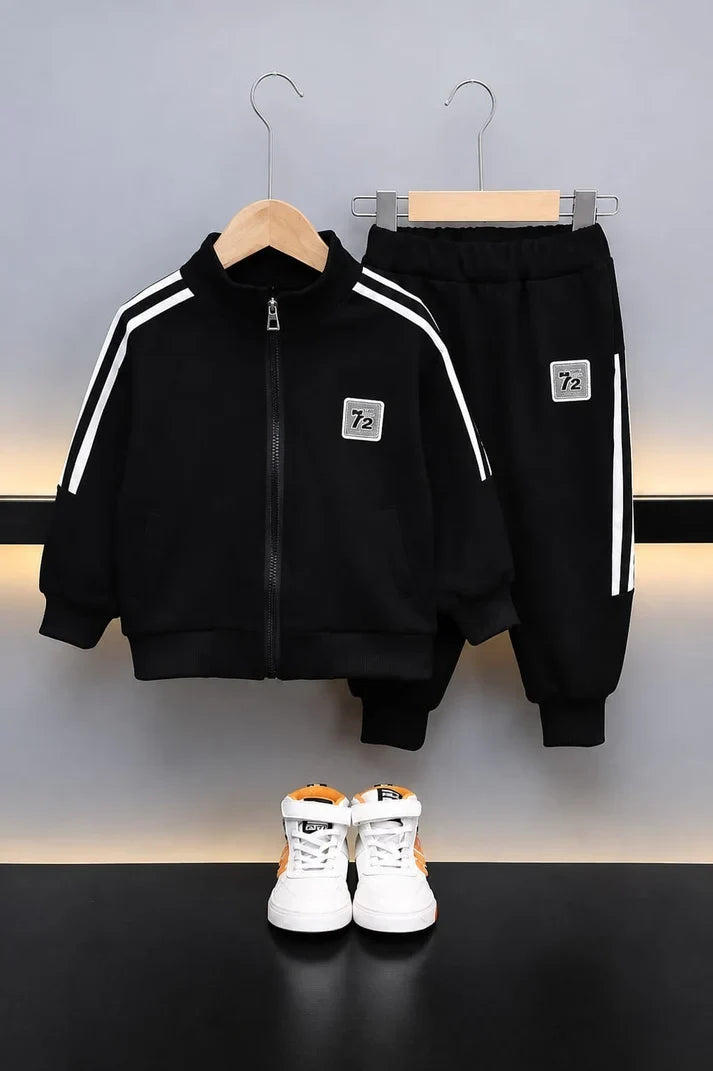 A two-piece kids' tracksuit set in black, featuring a zip-up jacket with white shoulder stripes and matching sweatpants, hanging on wooden hangers above a pair of white sneakers with orange accents.