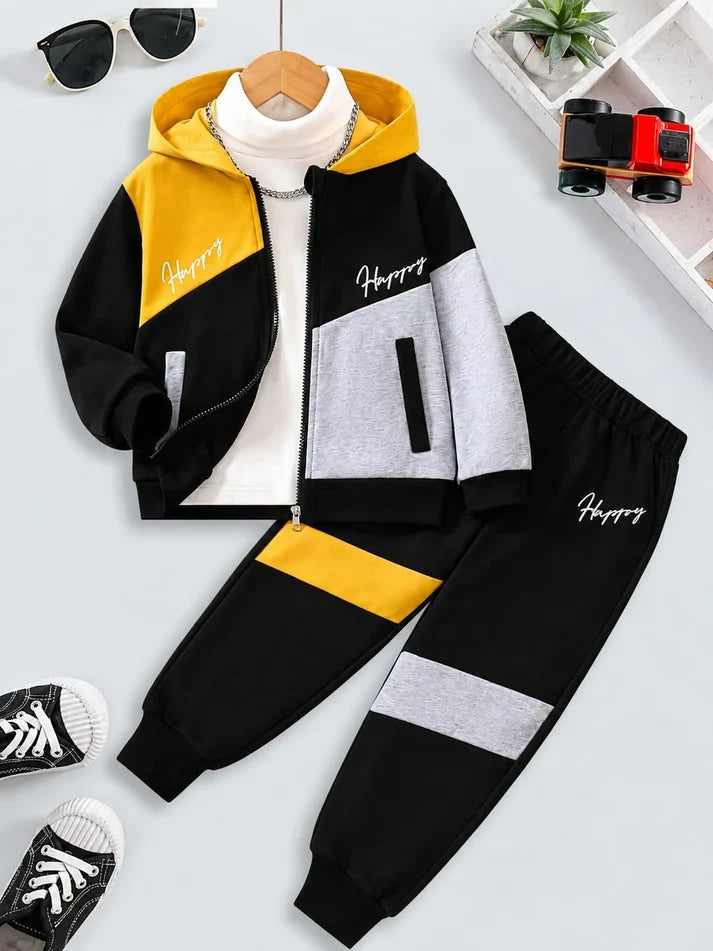 A children's two-piece tracksuit set laid out on a light gray background, featuring a black, yellow, and gray zip-up hoodie jacket with a white turtleneck underneath, paired with matching black jogger pants with gray and yellow color-block panels. Decorative props including sunglasses, a small plant, toy binoculars, and sneakers are arranged around the outfit.
