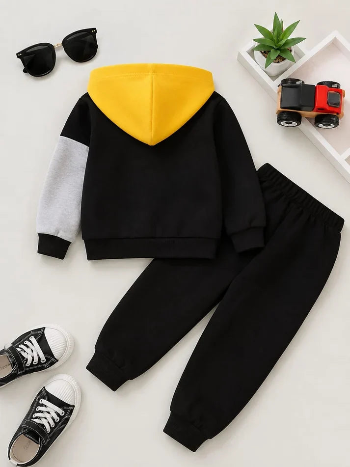 A children's two-piece clothing set laid flat on a light background, featuring a black hoodie with a bright yellow hood and a light gray sleeve, paired with matching black jogger pants. Styled with black sneakers, sunglasses, a small potted plant, and a toy truck.