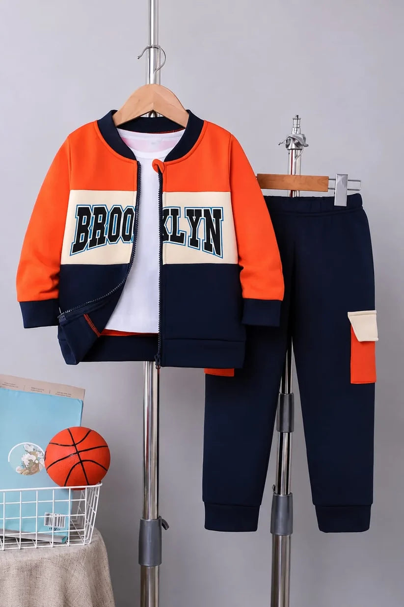 A children's two-piece tracksuit displayed on hangers against a light gray wall. The set includes an orange and navy blue zip-up jacket with 'BROOKLYN' across the chest, a white shirt underneath, and matching navy blue jogger pants with a side pocket detail.