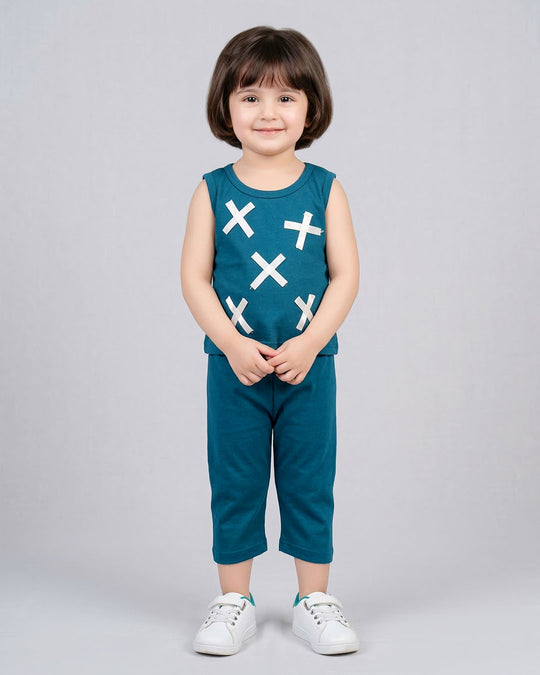 A young child with short brown hair stands against a light gray studio background wearing a sleeveless teal top with white X patterns and matching teal pants, paired with white sneakers.