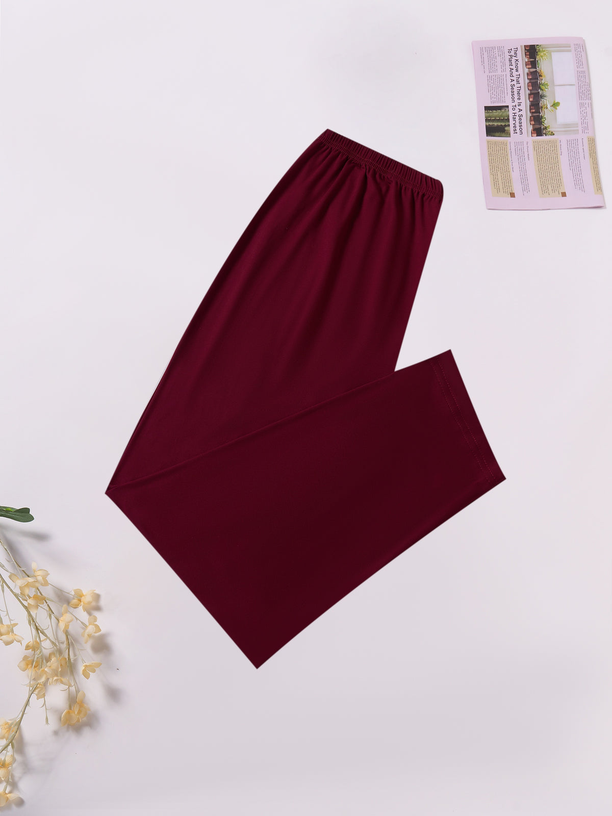 A pair of solid burgundy wide-leg lounge pants laid flat on a light gray background, styled with a small booklet in the top right and dried flowers in the bottom left.