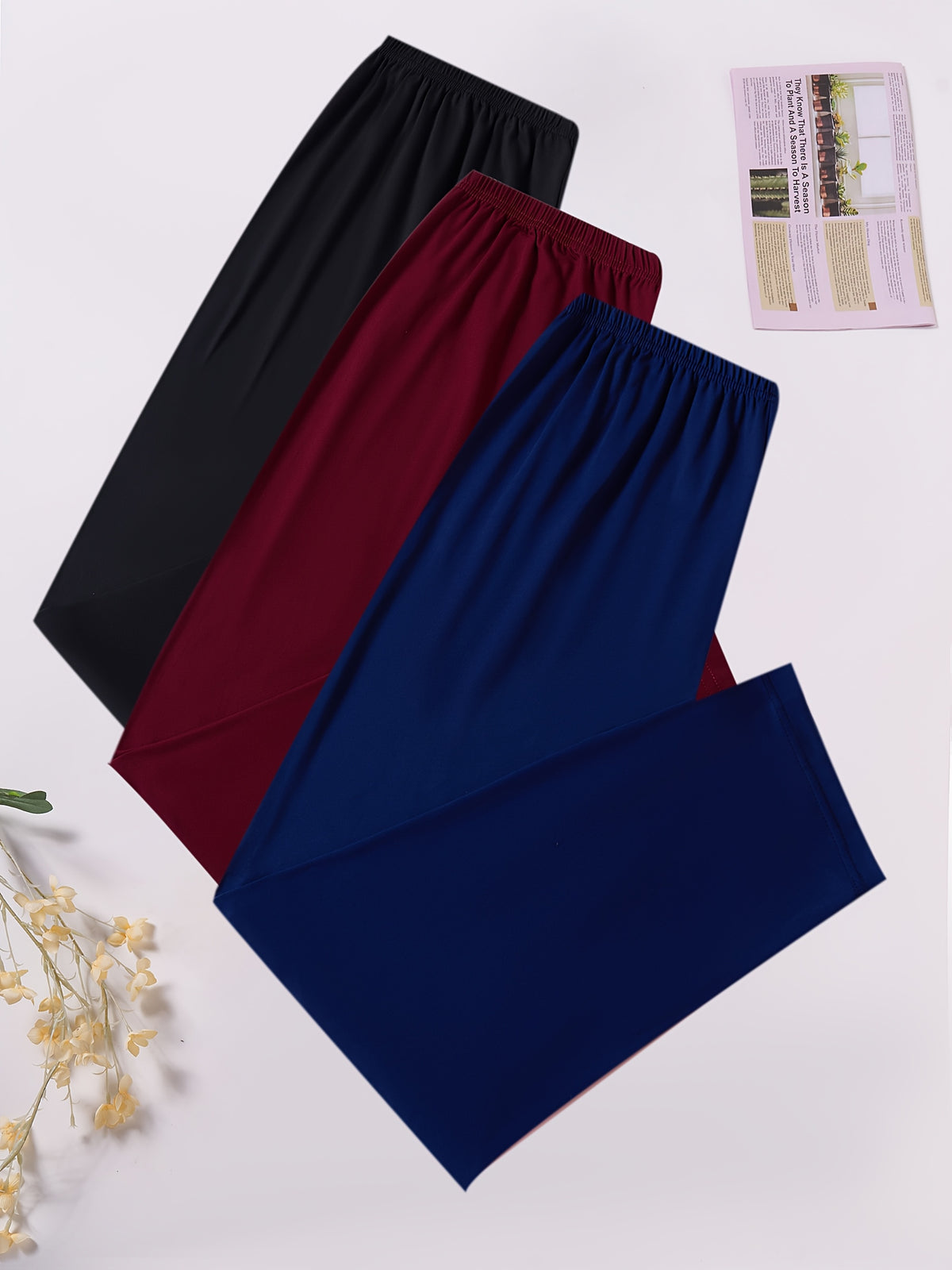 Three pairs of wide-leg lounge pants or pajama pants in black, burgundy, and navy blue, arranged diagonally on a white background with decorative dried flowers and a small magazine.