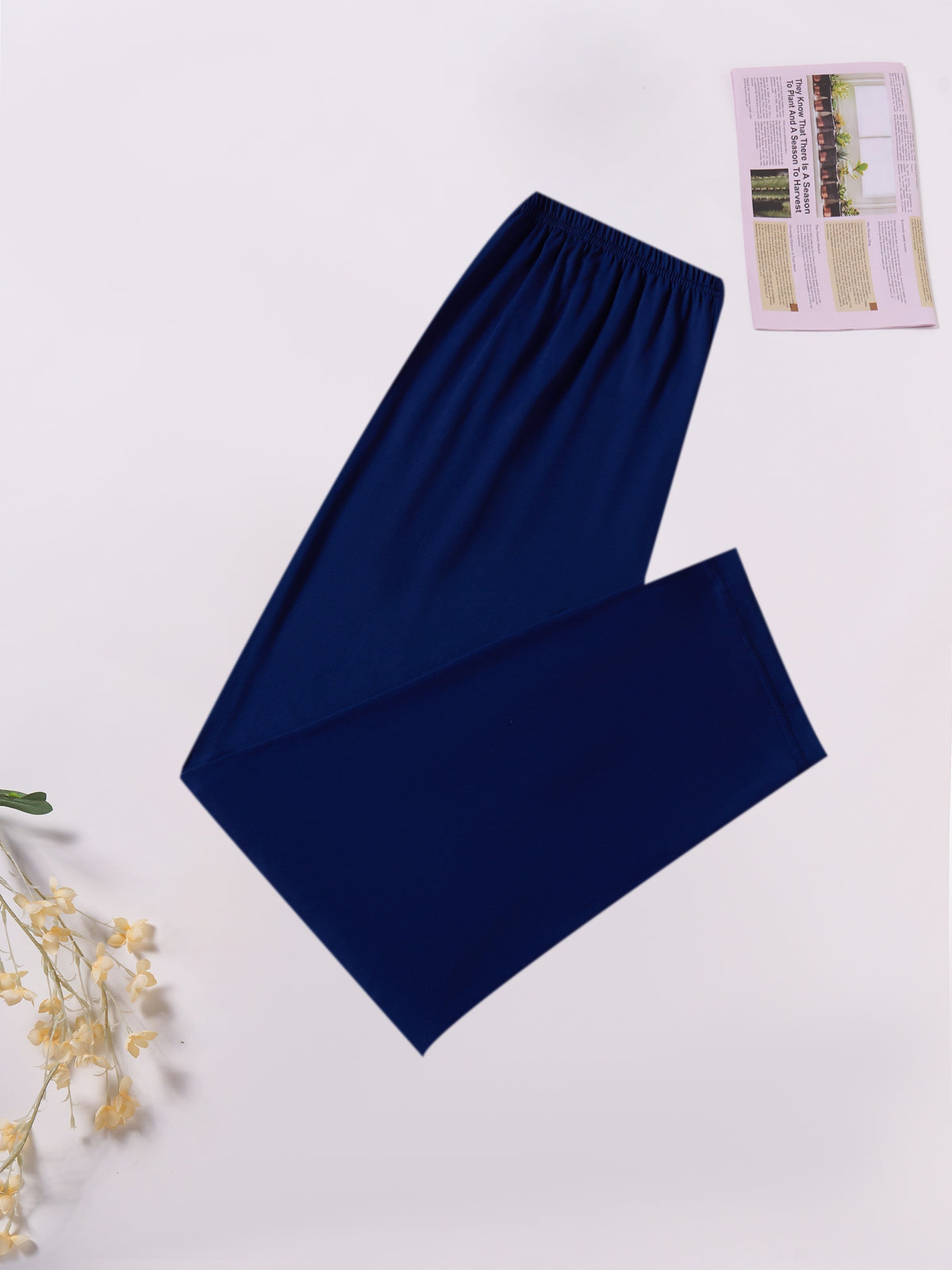 A pair of solid navy blue wide-leg lounge pants or pajama pants, folded neatly on a white background with decorative dried flowers and a small magazine page in the corners.