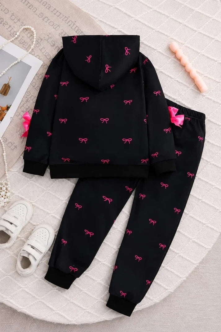 A two-piece kids' clothing set consisting of a black hooded sweatshirt and matching jogger pants, both decorated with a small pink bow pattern. The set is displayed in a styled flat-lay arrangement on a textured white blanket with decorative props including white sneakers, a pearl necklace, and a pink candle.