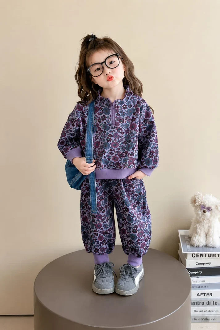 A young girl with glasses stands on a round pedestal wearing a matching purple and black floral print zip-up hoodie and jogger pants set, holding a blue shoulder bag in a studio setting.