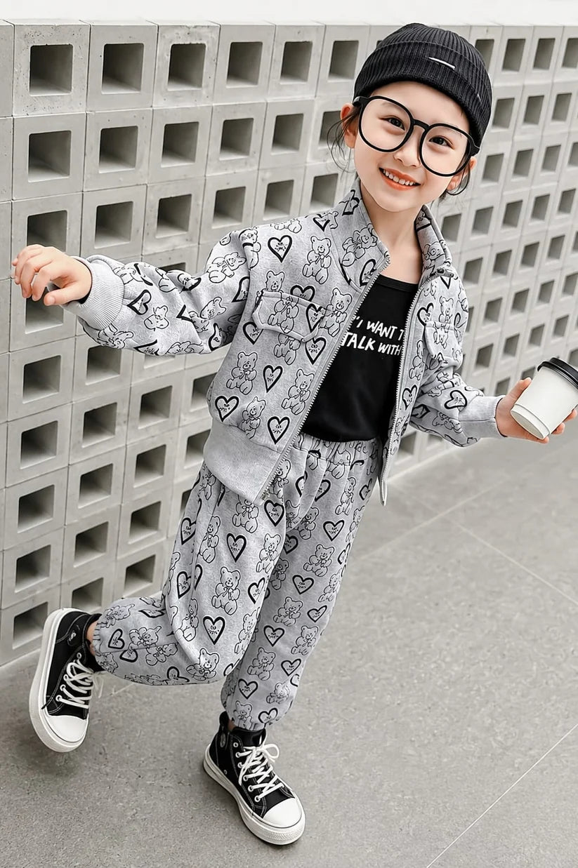 A young girl wearing a light gray two-piece tracksuit with black heart and cartoon patterns, a black graphic t-shirt, black sneakers, and a black beanie, posing in front of a gray concrete block wall.