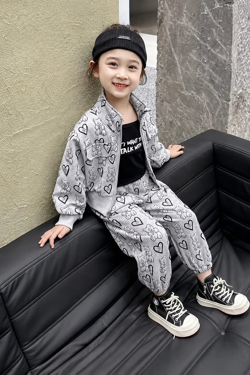 A young girl sitting on a black leather couch wearing a light gray two-piece tracksuit with black heart and cartoon patterns, a black graphic t-shirt, a black beanie, and black sneakers with white soles.