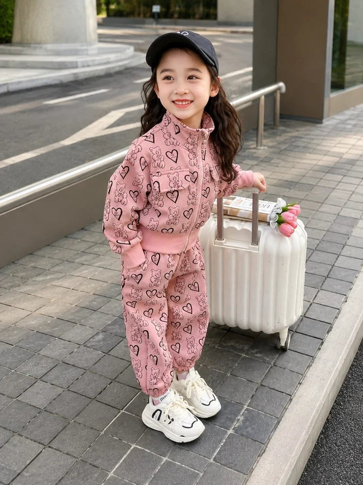 A young girl stands on a paved outdoor walkway next to a white suitcase, wearing a pink two-piece tracksuit with a black heart pattern, white sneakers, and a black cap.