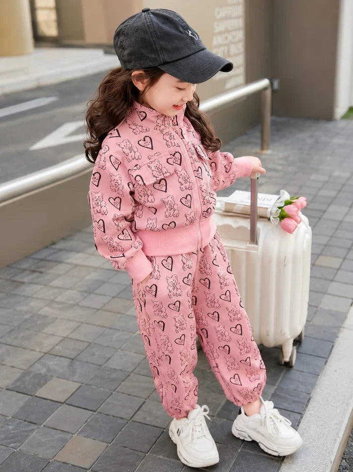 A young girl wearing a pink heart-patterned zip-up jacket and matching jogger pants, standing next to a white suitcase on a paved outdoor walkway.