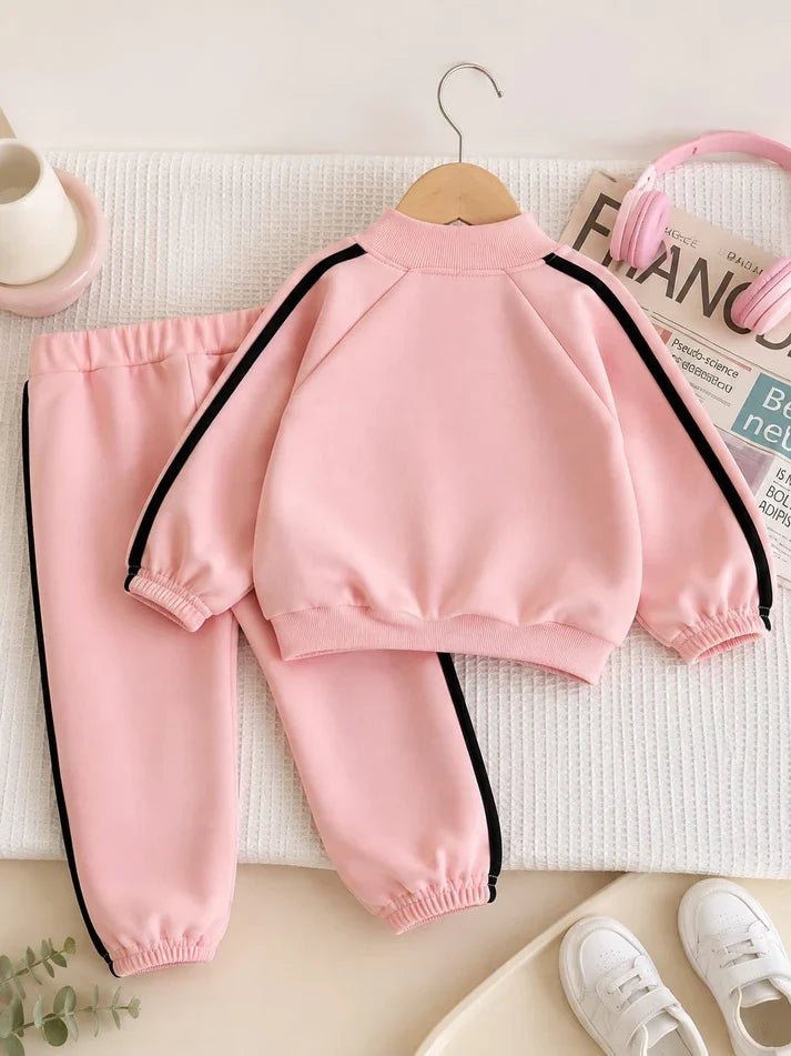 A two-piece children's tracksuit in light pink, featuring a mock-neck long-sleeve top with black side stripes and matching jogger pants, styled on a textured white blanket with props including headphones, a magazine, and white sneakers.