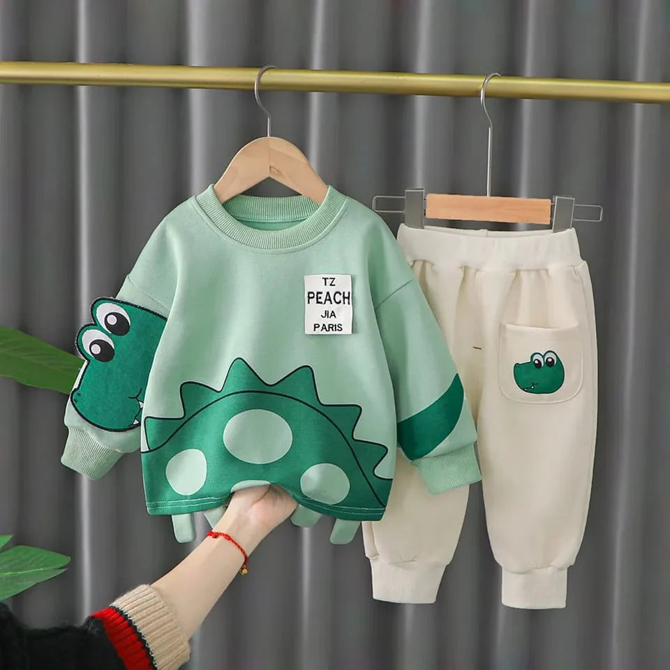 Children's green dinosaur-themed sweater and beige pants on hangers against a gray curtain background.
