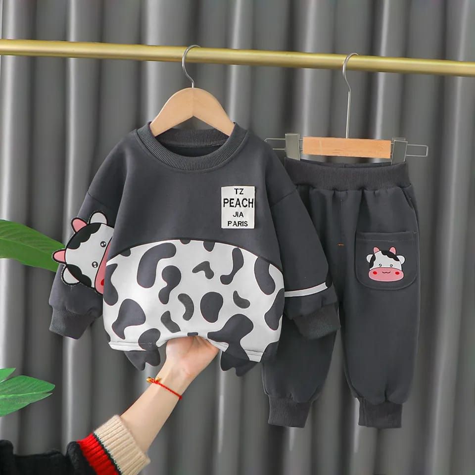 A children's two-piece sweatshirt and pants set hanging on wooden hangers against a gray curtain backdrop. The set is dark gray, featuring a cow-print pattern across the lower half of the sweatshirt and a matching cow graphic on the pants pocket.