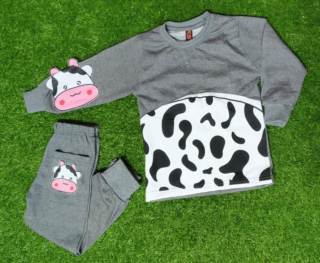A children's two-piece pajama set laid flat on green artificial grass. The set includes a long-sleeve gray top with a black-and-white cow print panel across the lower half and a cartoon cow face patch on the left sleeve, plus matching gray pants with a matching cow face patch on the leg.