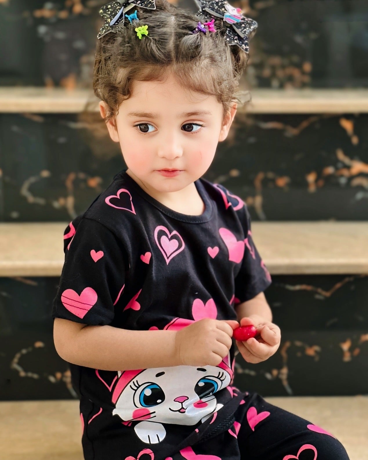 Child wearing a black shirt with pink hearts and a cat design, sitting on steps.