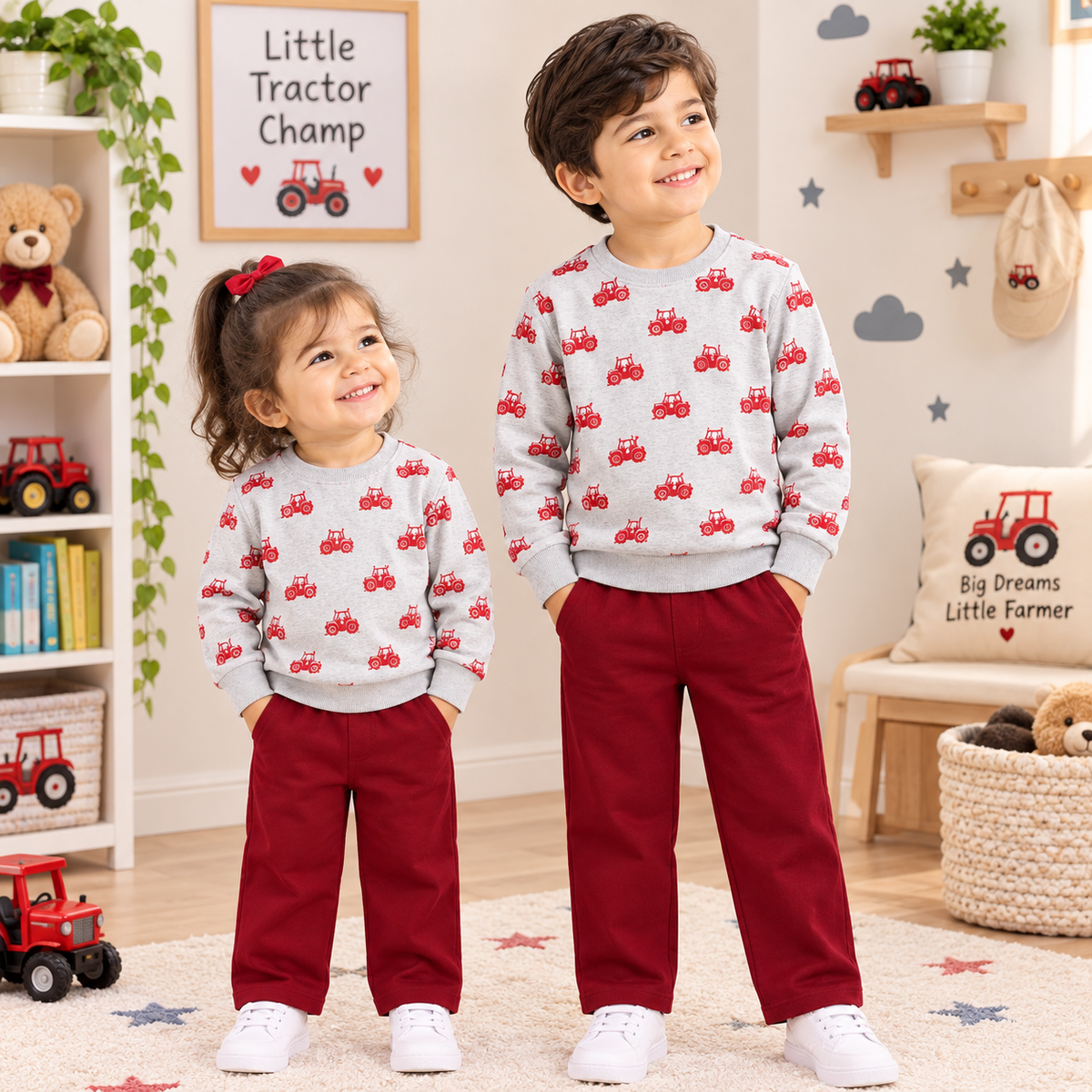 A young boy and girl stand in a child-friendly room wearing matching light gray sweatshirts with a red tractor print and dark red pants. The boy wears white sneakers, and the girl wears white sneakers with a red bow in her hair.
