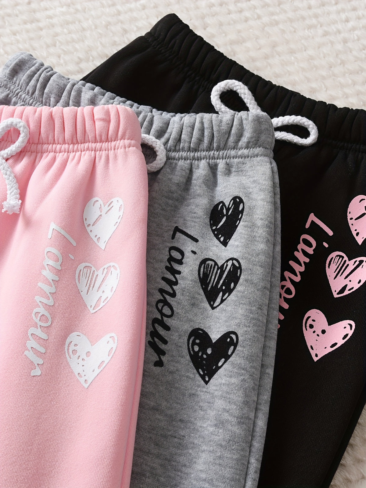 Three pairs of children's sweatpants in pink, gray, and black, laid out side-by-side. Each pair features an elastic waistband with white drawstrings and a graphic print of hearts and the word 'L'Amour' in a matching color.