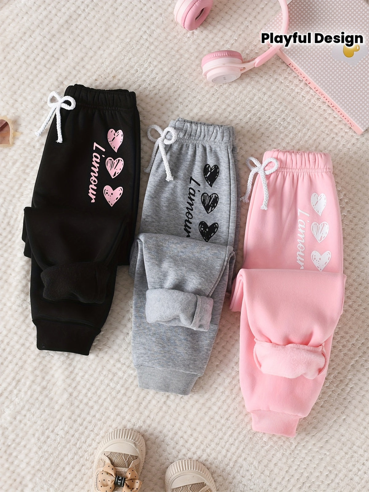 Three pairs of children's jogger sweatpants laid flat on a textured cream fabric: black with pink hearts and white text, gray with black hearts and white text, and pink with white hearts and text. Pink headphones and a small notebook are partly visible at the top, and beige shoes appear at the bottom.