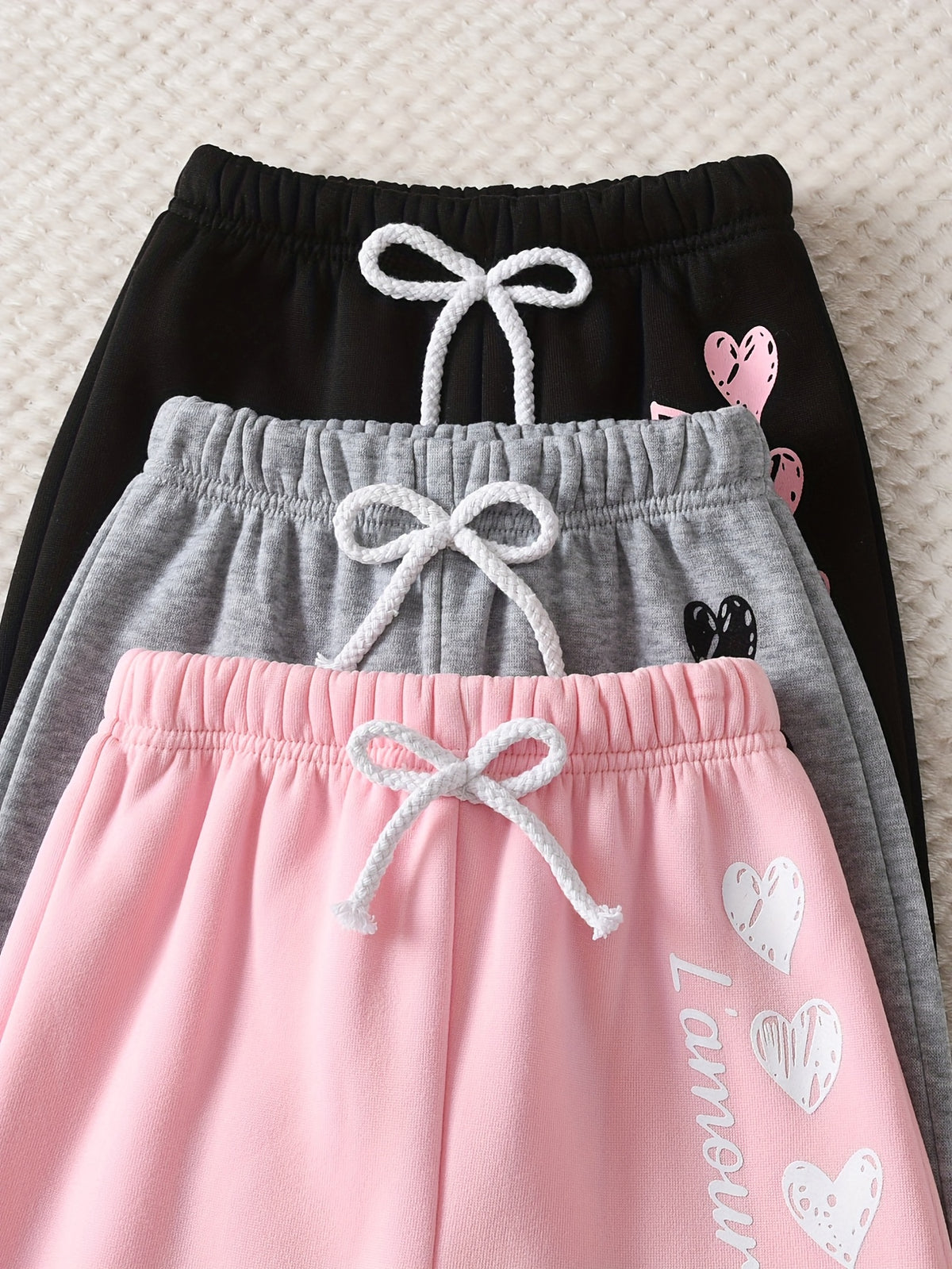 Three pairs of girls' drawstring sweatpants stacked together on a textured cream background: black with white drawstring and pink heart graphics, gray with white drawstring and small black heart graphic, and pink with white drawstring and white heart graphics plus vertical text.