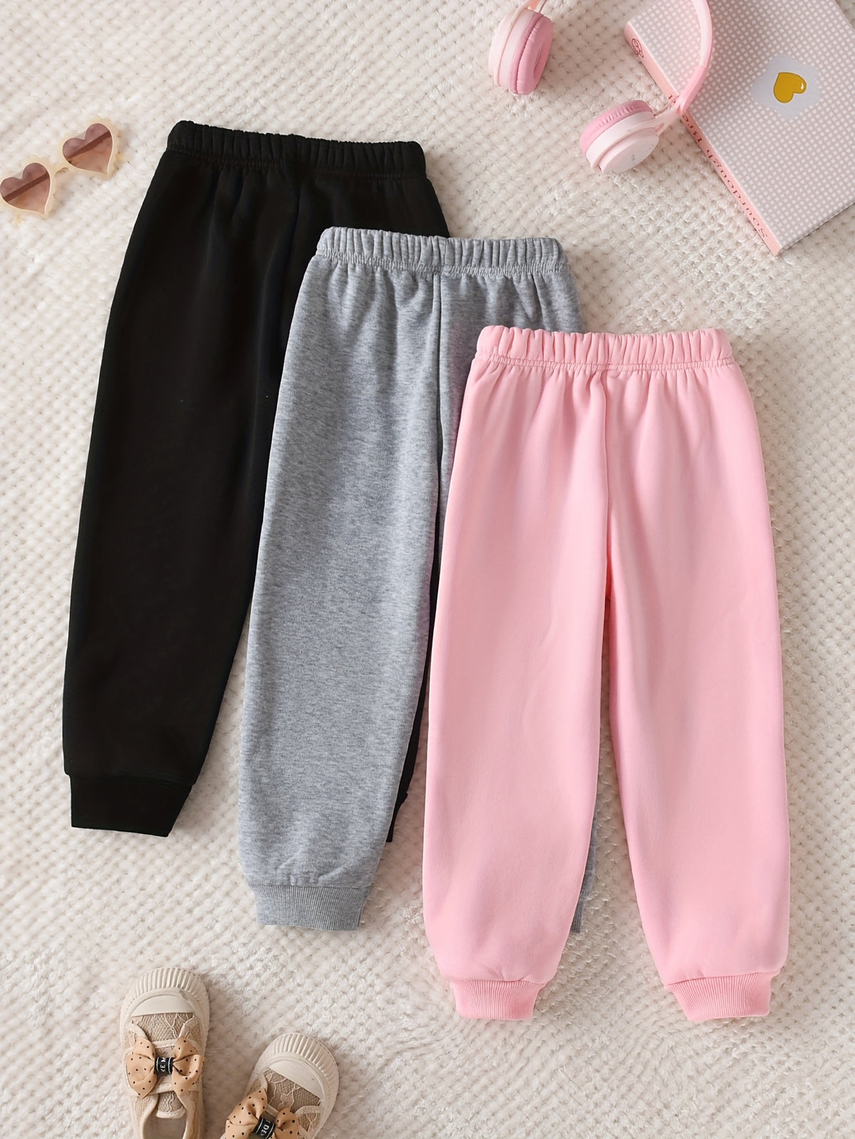 Three pairs of children's jogger sweatpants in black, gray, and pink, arranged in a flat lay on a textured cream blanket with small styling props including headphones, sunglasses, and baby shoes.