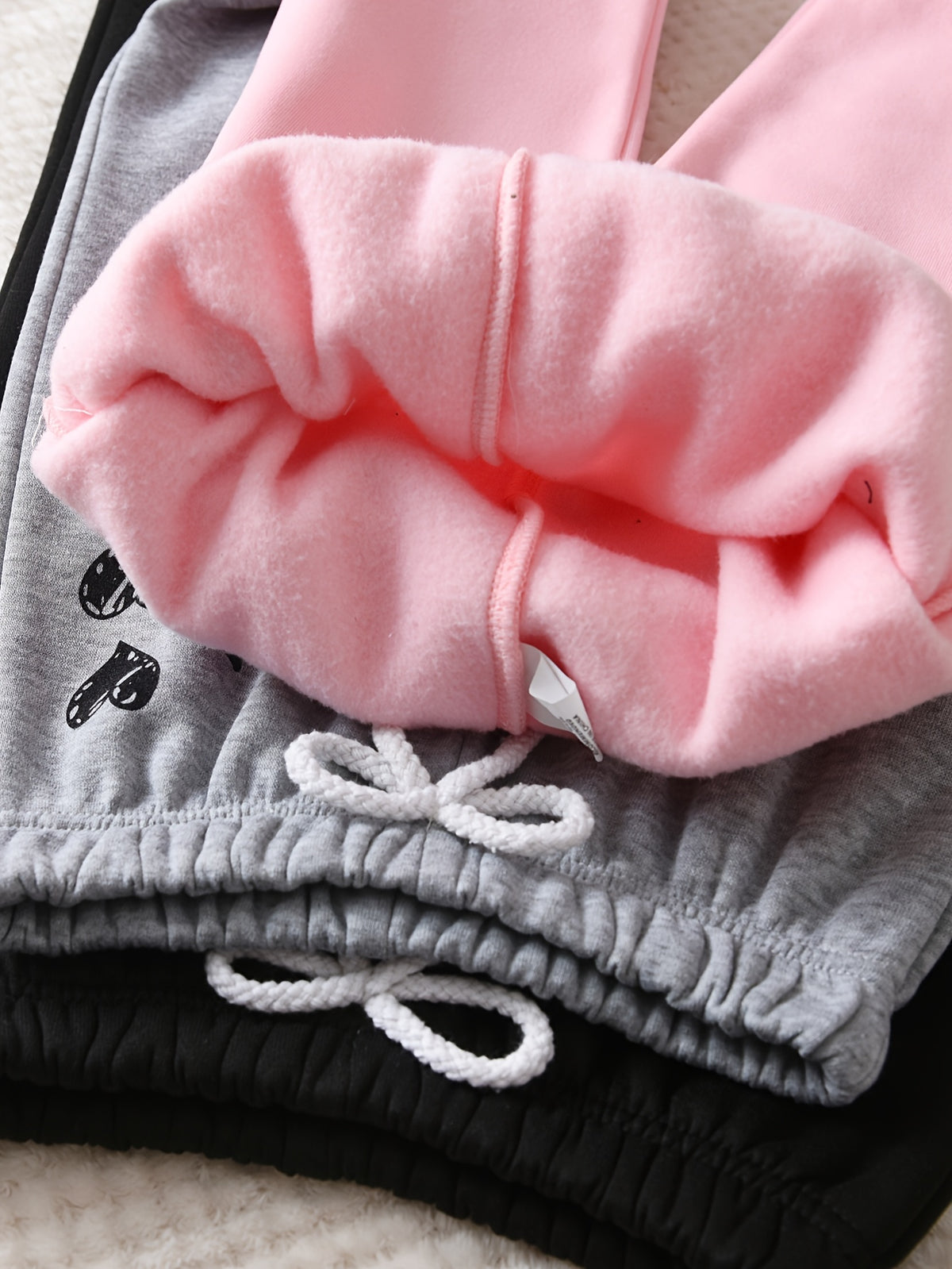 Close-up of folded sweatpants in three colorways: pink fleece lining, heather gray fleece lining, and black fleece lining, each with white drawstrings and a small black graphic on the leg.