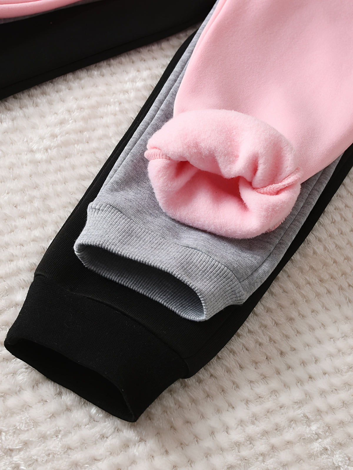 Close-up of a pair of sweatpants laid flat on a textured cream surface, showing a black outer layer and a soft pink inner layer with a folded cuff.