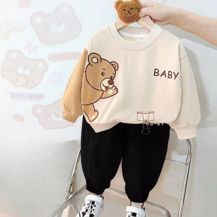 Children's outfit with beige sweater featuring a bear graphic and 'BABY' text, paired with black pants, on a white background.