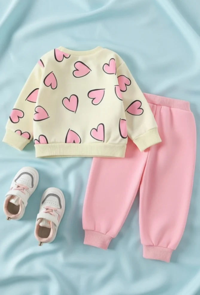 A toddler girl's two-piece outfit set featuring a cream sweatshirt with pink hearts and matching pink jogger pants, styled with a pair of white and pink sneakers on a light blue fabric background.