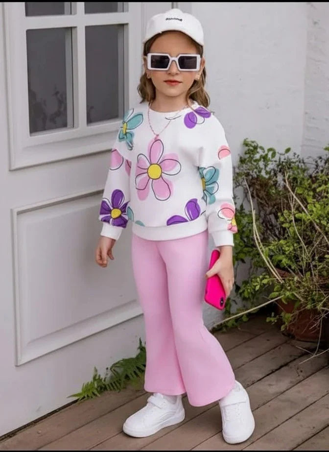 A young girl modeling a white sweatshirt with colorful floral prints, pink pants, white sneakers, a white cap, and sunglasses, standing on a wooden deck in front of a white wall.