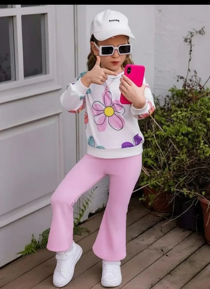 A young girl posing on a wooden deck wearing a white and pink outfit. The outfit consists of a white long-sleeve sweatshirt with a colorful flower graphic and pink pants.