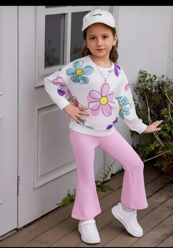 A young girl modeling a white sweatshirt with colorful daisy prints, pink flared pants, white sneakers, and a white baseball cap, standing on a wooden deck in front of a white door.