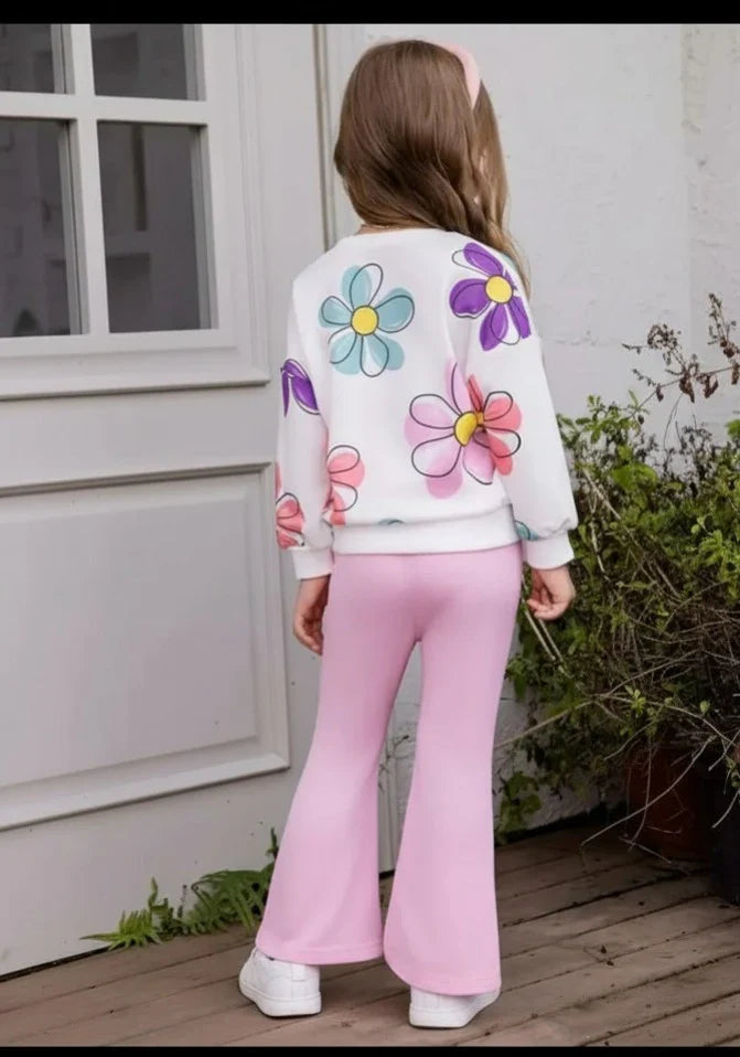 A young girl viewed from behind wearing a white sweatshirt with colorful floral appliques and light pink flared pants, standing on a wooden deck in front of a white house exterior.