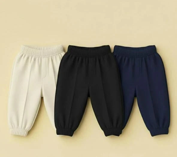 Three pairs of children's elastic-waist jogger pants in off-white, black, and navy blue, arranged side-by-side on a solid beige background.
