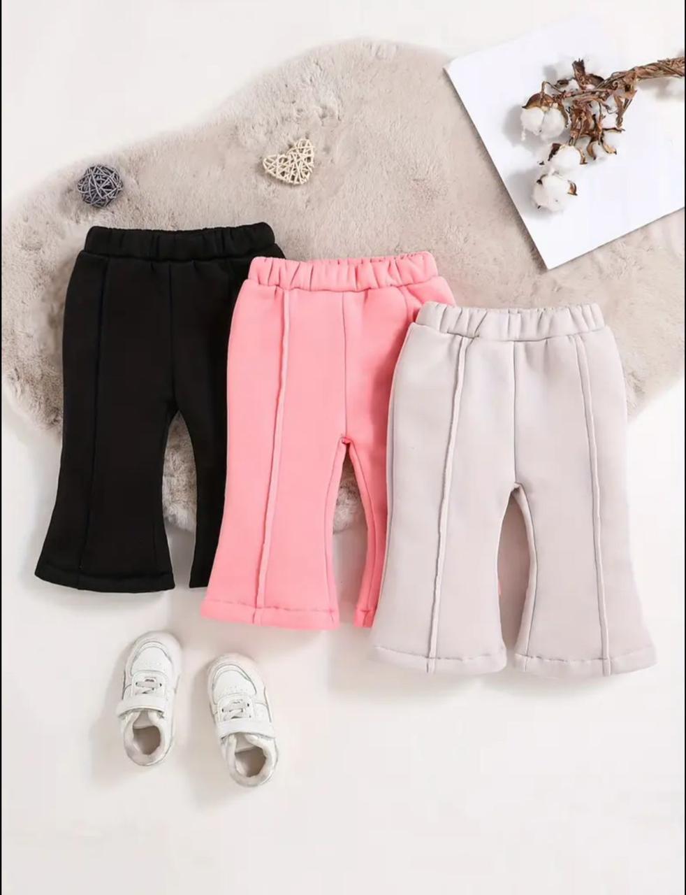 Three pairs of toddler girls' wide-leg sweatpants in black, pink, and light gray, styled in a flat-lay with white sneakers, a faux fur rug, and decorative props.