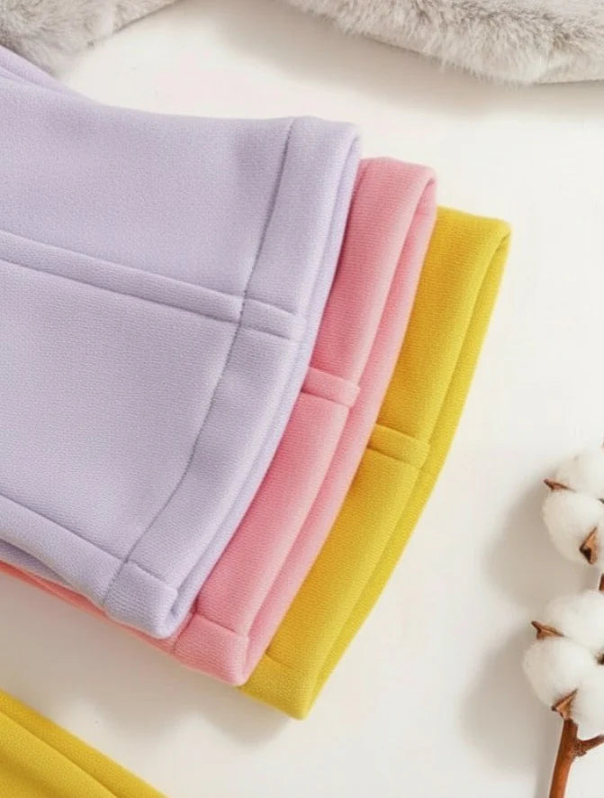 Three folded pairs of soft knit pants or sweatpants in pastel lavender, pink, and mustard yellow, arranged diagonally on a white surface with cotton stems and a fuzzy gray textile as props.