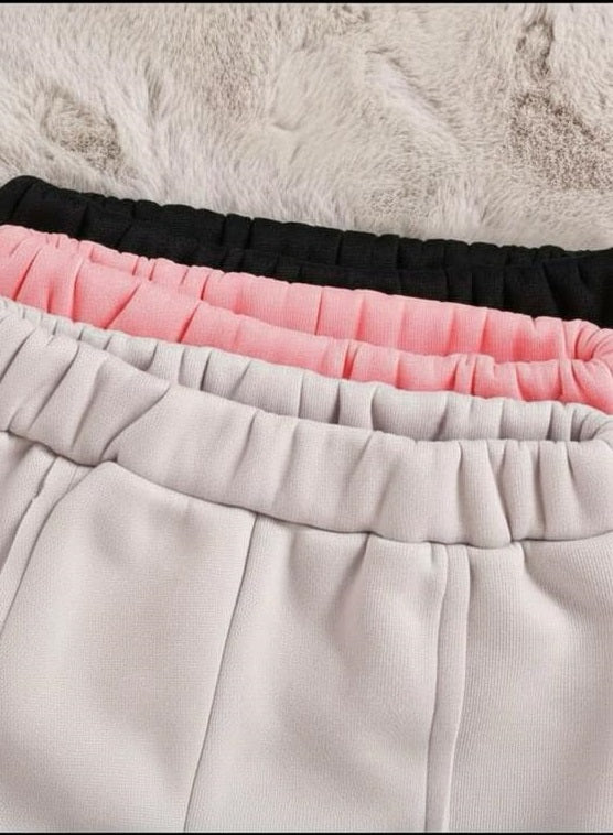 Close-up of three pairs of pleated sweatpants or shorts in black, pink, and light gray, stacked diagonally on a white faux fur surface.