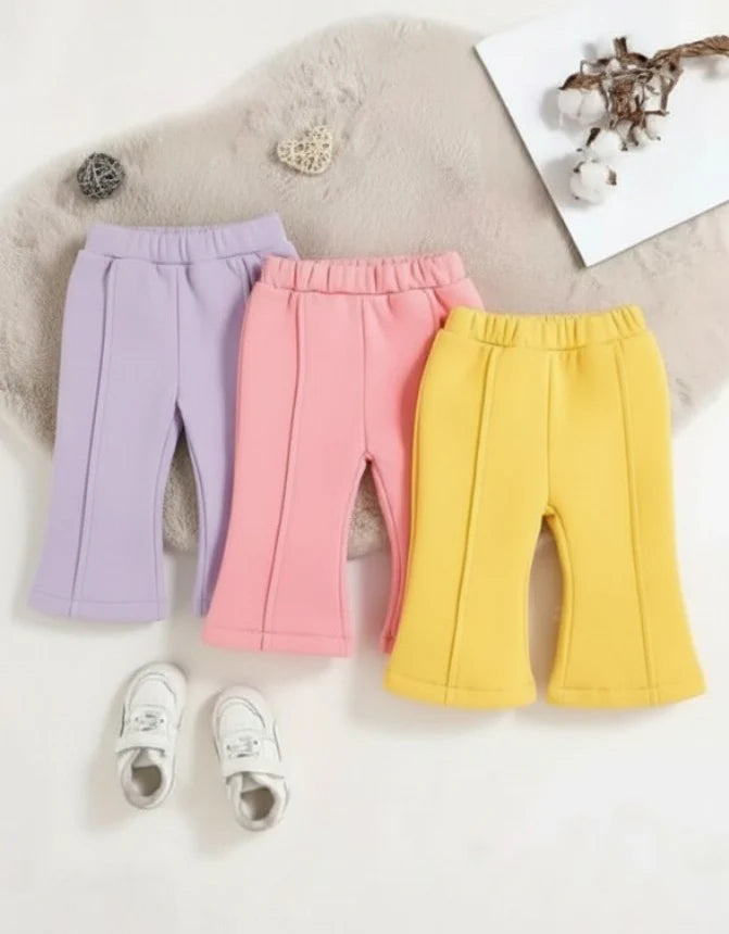 Three pairs of children's wide-leg sweatpants in pastel purple, pink, and yellow, arranged on a white surface with a beige faux-fur pad, small white shoes, and decorative props.