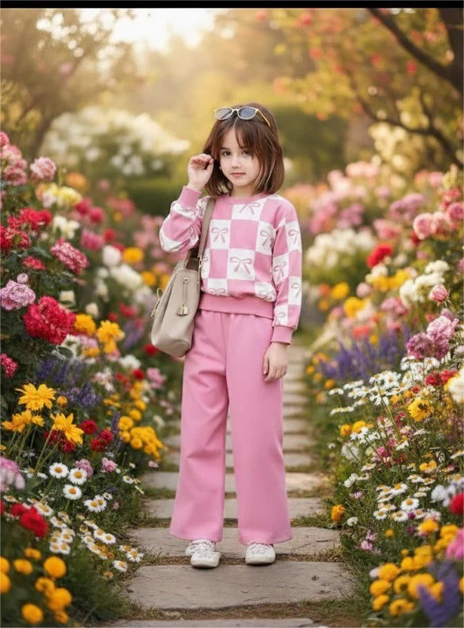 A young girl stands on a stone path in a flower garden, wearing a pink and white checkered sweatshirt, pink wide-leg pants, white sneakers, and carrying a beige shoulder bag.