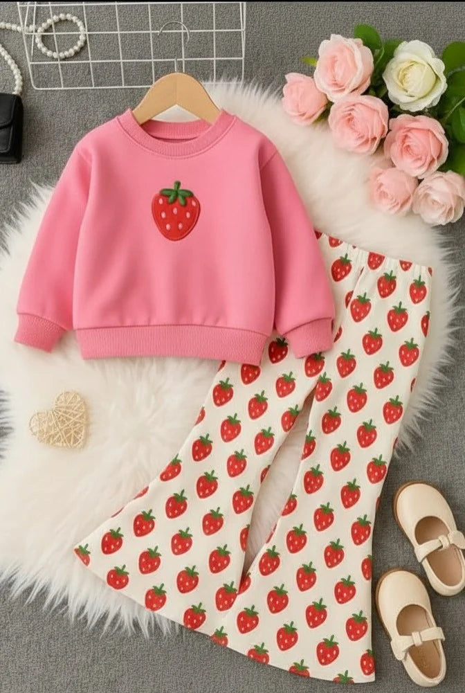 A children's two-piece outfit set featuring a pink sweatshirt with a strawberry graphic and matching cream pants with a strawberry print, styled on a white faux fur rug with decorative flowers and shoes.