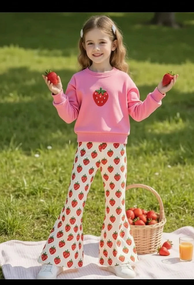 A young girl stands on a picnic blanket in a grassy park, wearing a pink sweatshirt with a strawberry graphic and white flared pants covered in a red strawberry print. She holds two strawberries, with a basket of strawberries and a glass of juice beside her.