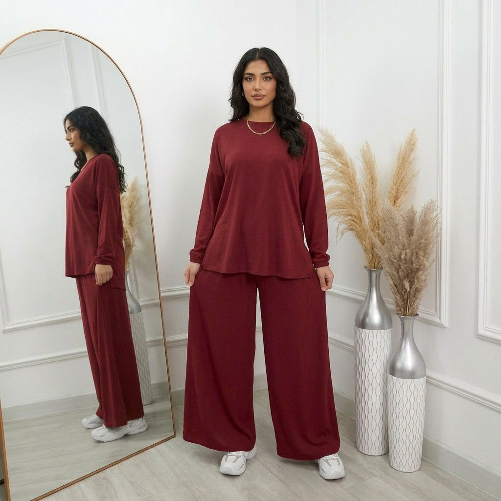 A woman modeling a matching burgundy two-piece lounge set consisting of a long-sleeve top and wide-leg pants, standing in a bright room with a large arched mirror, pampas grass in vases, and white sneakers.