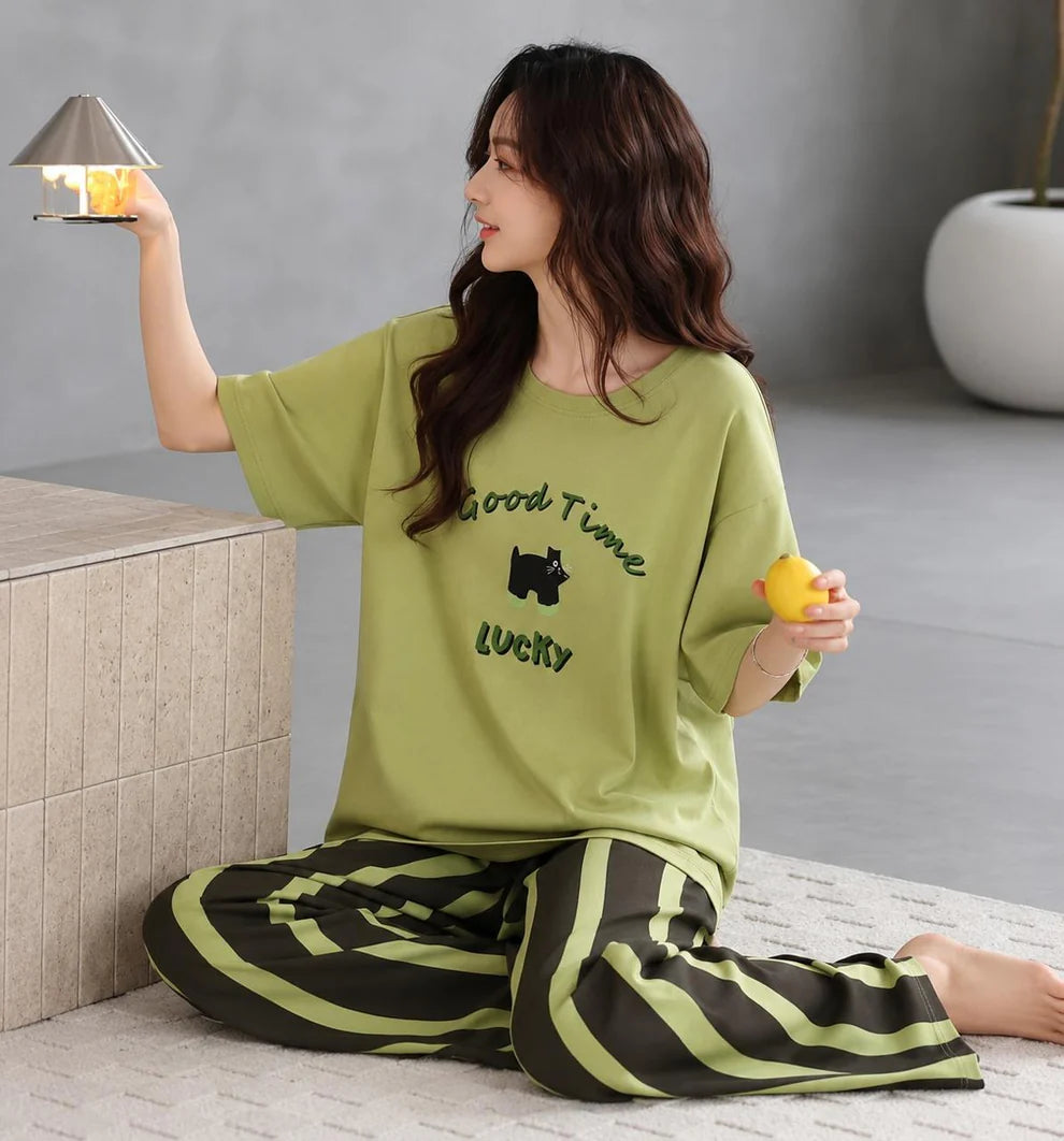 A woman sits on a patterned floor in a modern room, wearing a light olive green short-sleeve pajama top with 'Good Time Lucky' and a bear graphic, paired with matching green and black striped pants. She holds a small yellow duck toy and a lit lamp.